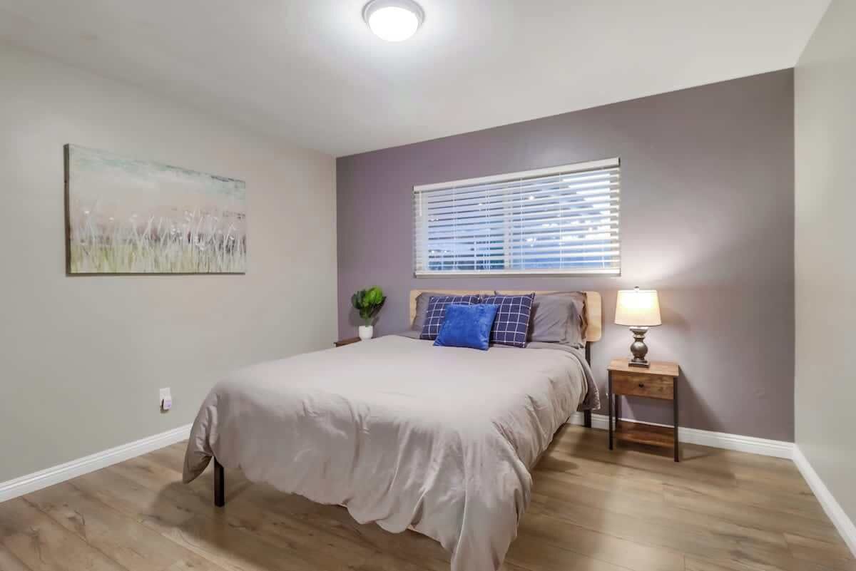 A bedroom with a bed, a nightstand, a lamp, a window with blinds, and a painting on the wall.