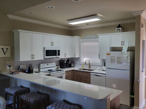 A kitchen with white cabinets, a microwave, stove, and a refrigerator. There is a window with blinds and a sign above the refrigerator.