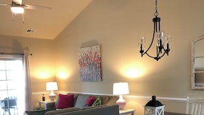 Una sala de estar acogedora con un sofá gris, una mesa de comedor de madera con sillas blancas, una araña y un cuadro en la pared.