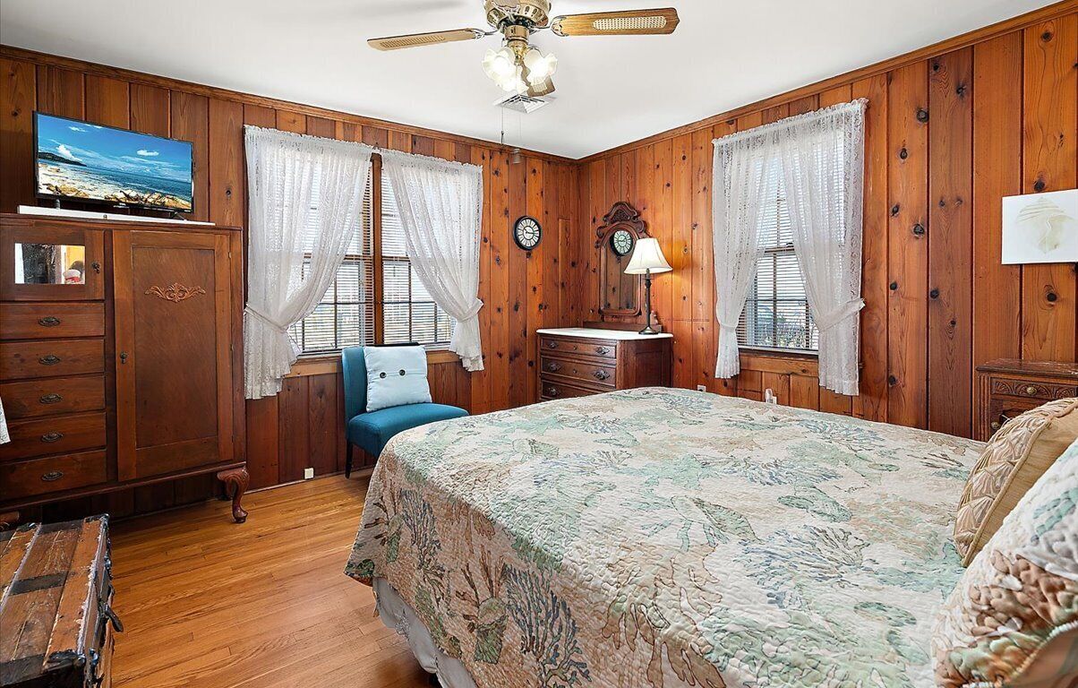 Una habitación con paneles de madera, una cama con colcha floral, una silla azul, un aparador, un televisor y un ventilador de techo.