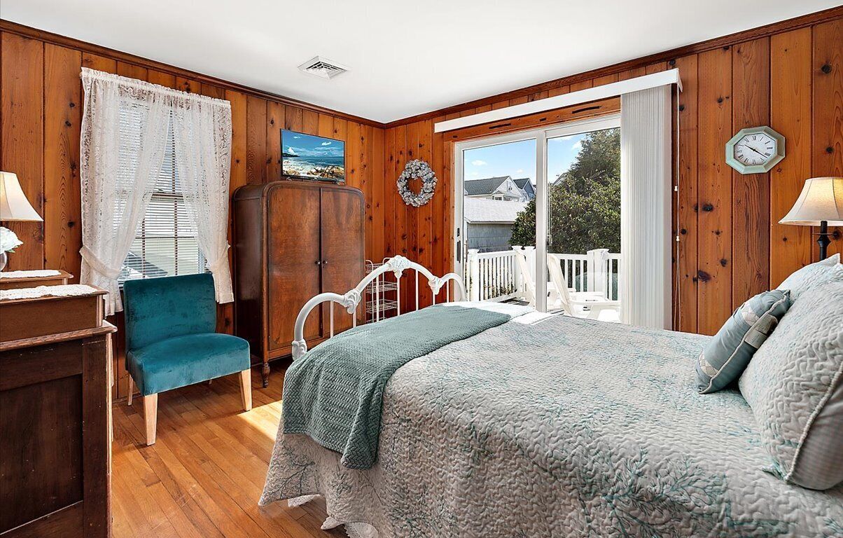 A bedroom with a bed, a chair, a wardrobe, a window with curtains, a door, and a view of the outside.