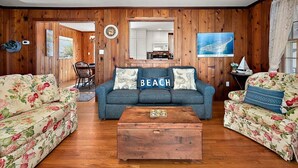 Un salon aux murs en bois, un canapé agrémenté d’un coussin orné du mot « PLAGE », une table basse et une vue sur la cuisine, visible à travers un miroir.