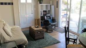 A living room with a sofa, a chair, a small table, a basket, and a sliding glass door leading to an outdoor area. - Beach Cottage - 4th from Ocean, Private Patio, Ocean View, now with A/C (San Diego)