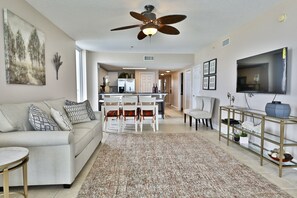 A spacious living room with a ceiling fan, a TV, a dining area with chairs, and a kitchen in the background.