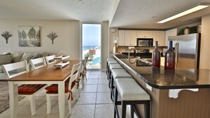 A modern kitchen with a large island, white cabinetry, and stainless steel appliances. There is a dining area with a wooden table and chairs, and a view of the ocean through a large window.