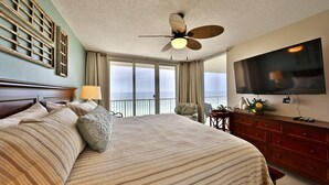 A hotel room with a large bed, a ceiling fan, a TV, a balcony with a view of the ocean, and a wooden dresser.