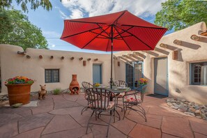 An outdoor patio with a red umbrella, stone flooring, rustic chairs, and potted plants.