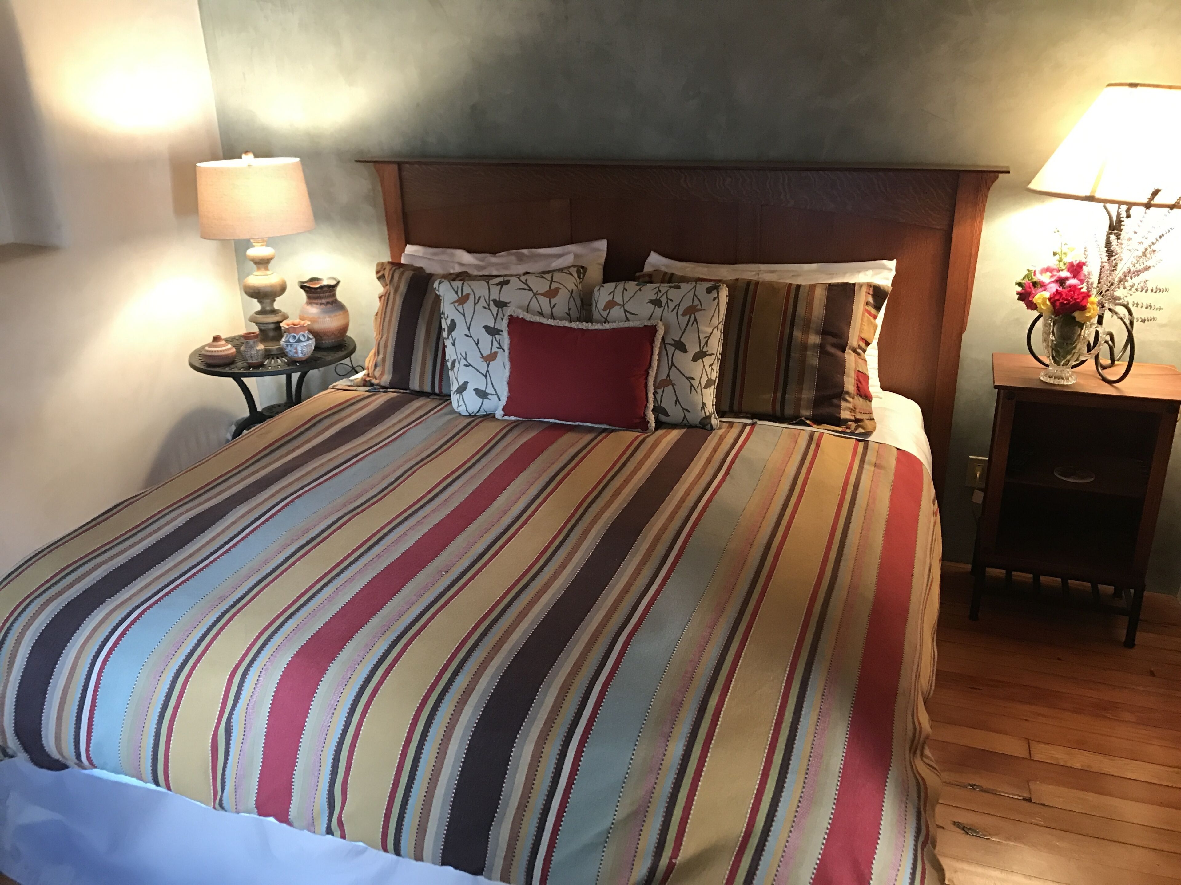 Una cama con una colcha a rayas, un cabecero de madera y varias almohadas. A la derecha, una mesita de noche con una lámpara y un jarrón con flores.