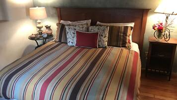 Una cama con una colcha a rayas, un cabecero de madera y varias almohadas. A la derecha, una mesita de noche con una lámpara y un jarrón con flores.