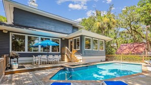 Uma casa com piscina, varanda coberta com cadeiras e guarda-sol, e deck com espreguiçadeiras.