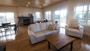 Un salon spacieux avec un plafond en bois, une cheminée en pierre, un canapé blanc, une table basse en bois et un fauteuil blanc.
