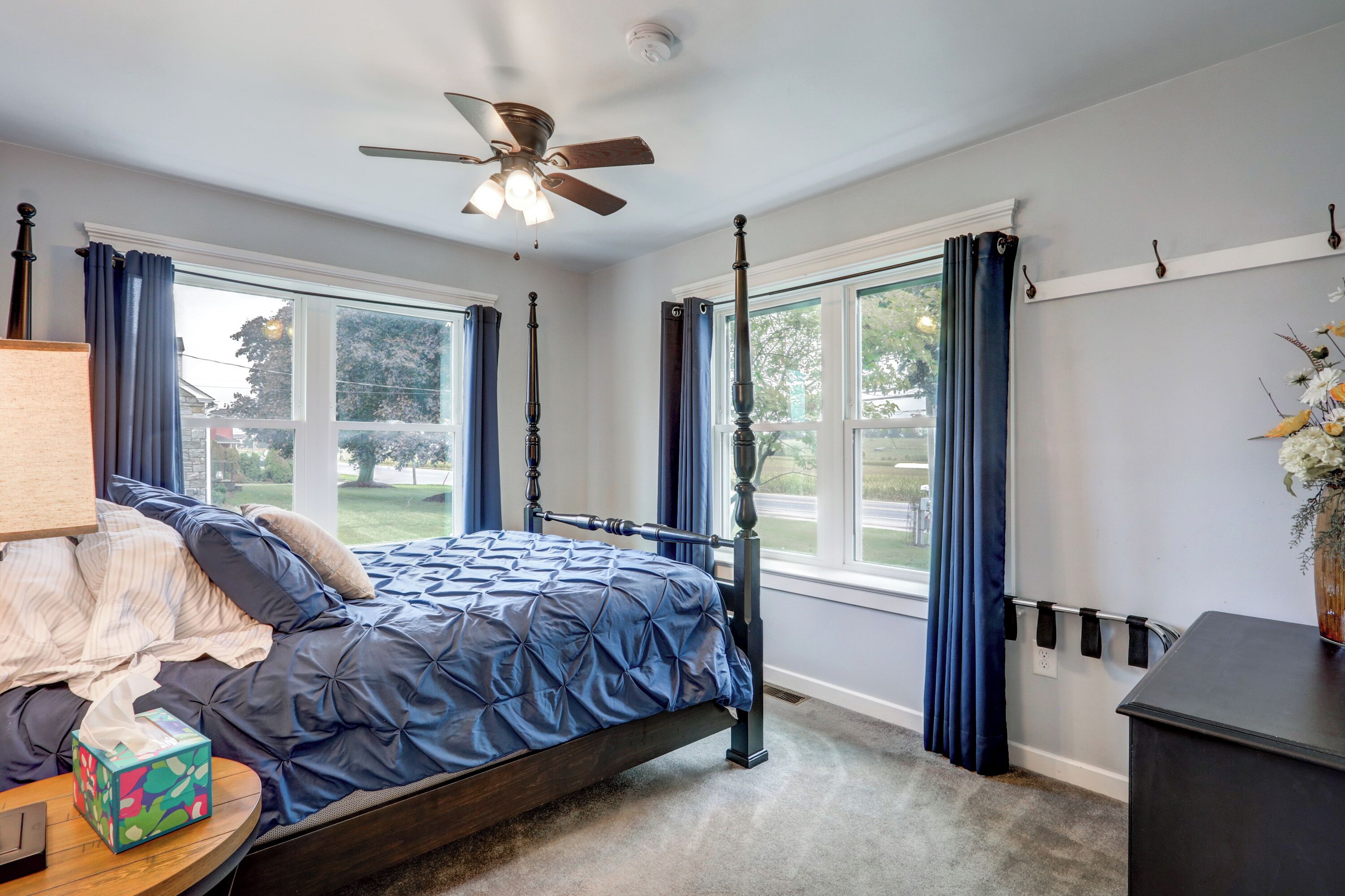 Quarto com cama de dossel, ventilador de teto, janelas amplas com cortinas e mesa de cabeceira com abajur e lenços de papel.