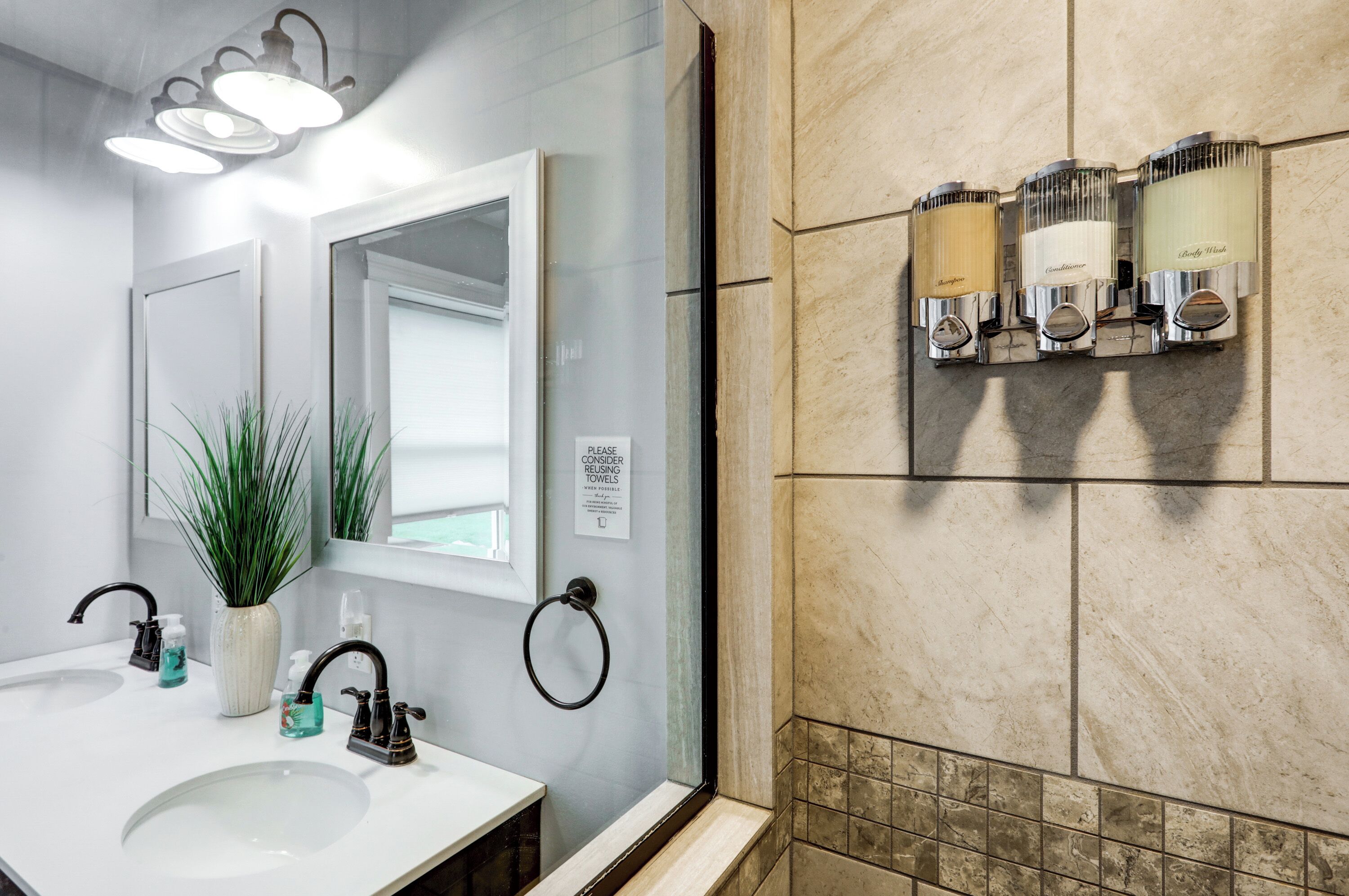 A modern bathroom with a white sink, two faucets, a mirror, a potted plant, and a glass shower enclosure.