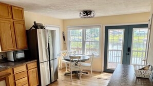 Une cuisine avec des meubles en bois, un réfrigérateur, une table à manger avec des chaises et une grande fenêtre donnant sur un paysage verdoyant.