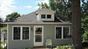 Une petite maison de plain-pied avec un porche à l’avant, un petit garage attenant et un arbre en arrière-plan.