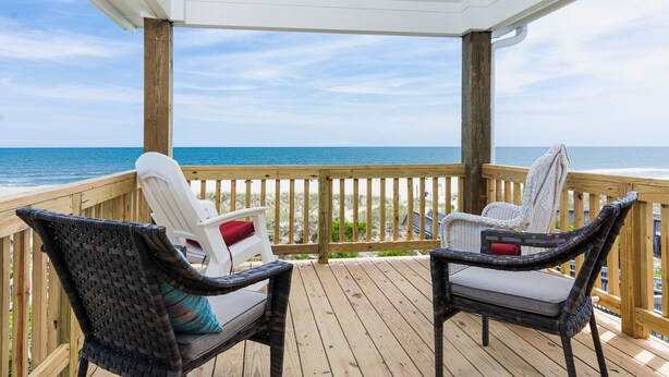 Une terrasse en bois avec deux fauteuils en osier et une chaise blanche, donnant sur une plage et l’océan.