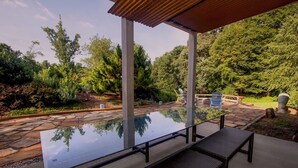 Une terrasse couverte avec une table en verre et des bancs, entourée de chemins de pierre et d’une végétation luxuriante.