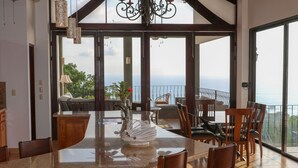 A spacious kitchen with a large island, dining area with chairs, and a view of the ocean through large windows. - Tropical Villa with Spectacular Views!   (Dominical)