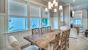 A dining area with a wooden table and chairs, large windows with blinds, a chandelier, and a television mounted on the wall. - Lavish Coastal Getaway w/Media Room | 3 Min to Beach | Steps to Pool | 6 Bikes (Inlet Beach)