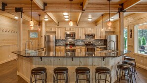 A spacious kitchen with wooden cabinets, a central island with stools, stainless steel appliances, and pendant lighting. - Home Theater, Hot tub & Huge Covered Patio! Stunning Home 2 Miles from Town! (Leavenworth)