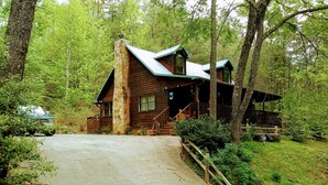 Une cabane rustique en rondins de bois, dotée d’une cheminée en pierre, d’un porche et d’une allée en béton menant à l’entrée.