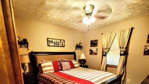 Une chambre à coucher comprenant un lit, un ventilateur de plafond, une table de chevet, une lampe, une fenêtre avec des rideaux et des œuvres d’art murales.