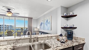 A kitchen with a double sink, granite countertop, and a ceiling fan. There are chairs and a table outside the window, and artwork on the wall.