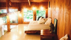 Ein Schlafzimmer einer Holzhütte mit einem Bett, Nachttisch und einem Fenster mit Blick auf Grünflächen.