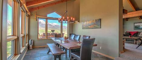A dining area with a long wooden table, benches, and chairs. There are large windows with a view of mountains.