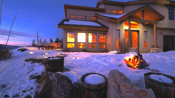Une maison moderne avec de grandes fenêtres, un foyer extérieur et un sol recouvert de neige.