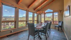 Un coin repas avec une table et des chaises, de grandes fenêtres offrant une vue sur les montagnes, des poutres en bois au plafond et un lustre.