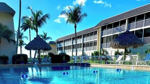 A clear blue pool with a lifeguard chair, palm trees, and a building with balconies.