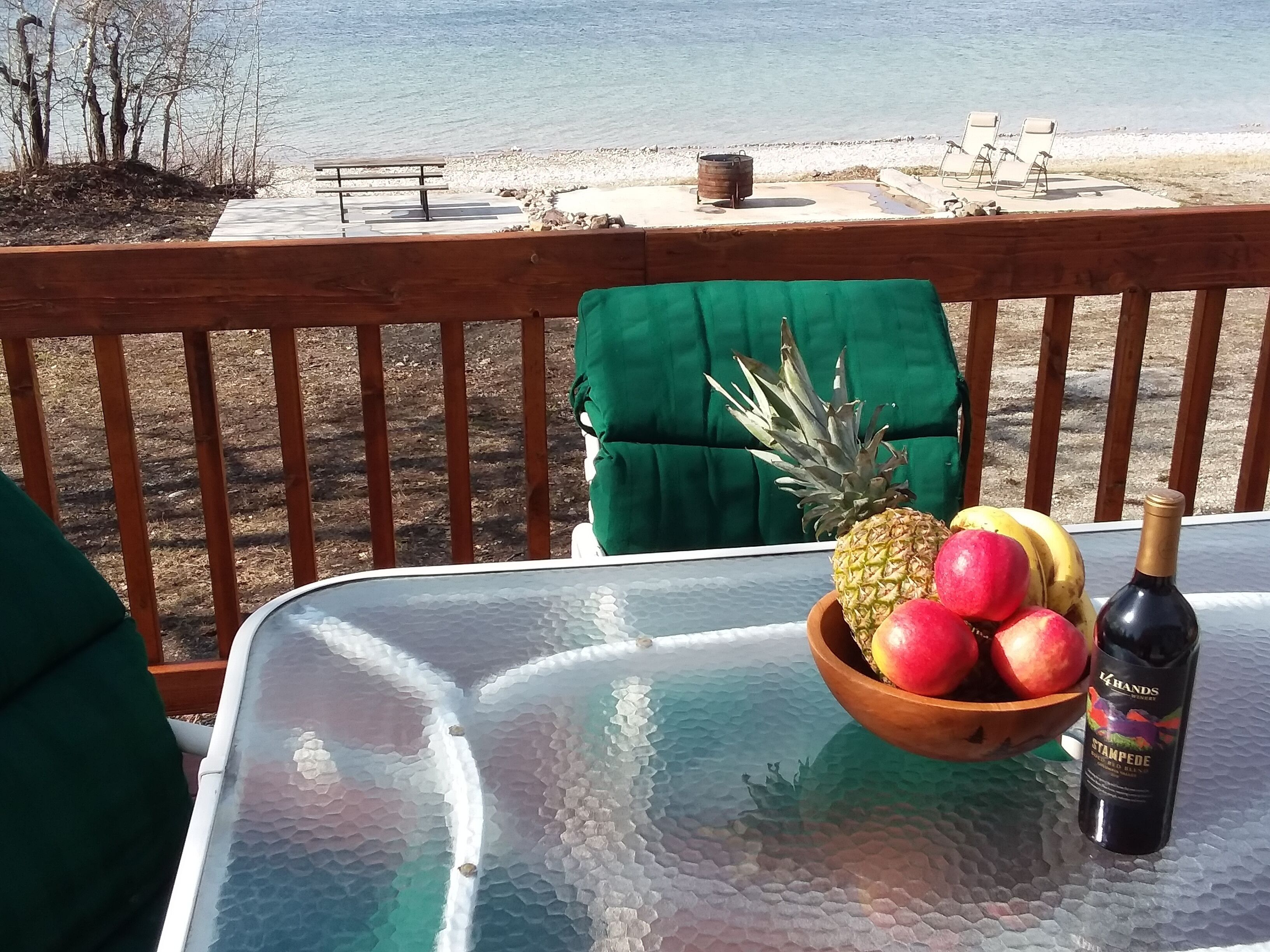 Una mesa con cubierta de vidrio, un cuenco de madera con fruta y una botella de vino en un balcón con vistas a la playa.
