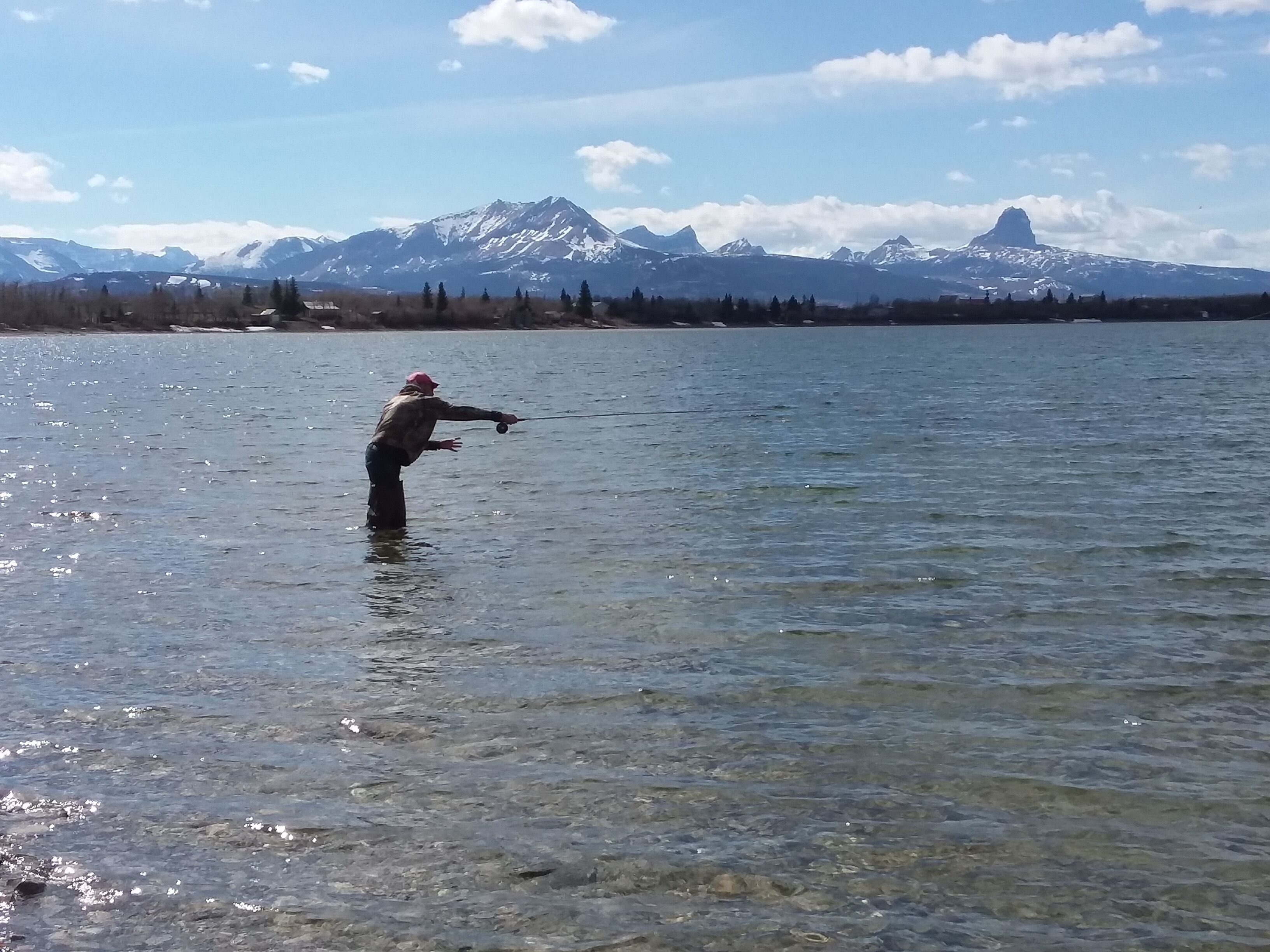 Une personne est en train de pêcher dans un lac aux eaux claires, avec des montagnes en arrière-plan.