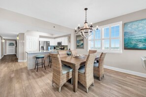A dining area with a wooden table and wicker chairs, a chandelier, and a view of the ocean through large windows.