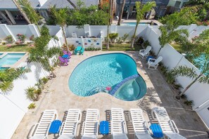 A swimming pool with a unique curved design, surrounded by white walls and palm trees. There are lounge chairs and a small deck area with a table and chairs.
