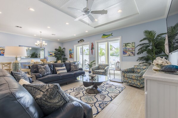A spacious living room with a dark blue sofa, patterned cushions, a glass coffee table, and a large potted plant.