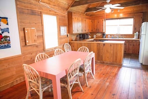 Interior de una cabaña de madera con comedor, cocina y un ventilador de techo.