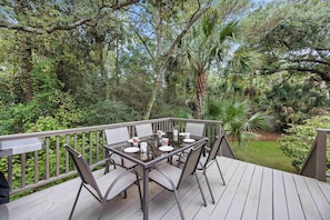 Un espacio de comedor al aire libre con una mesa de vidrio y sillas metálicas, rodeado de abundante vegetación y árboles.