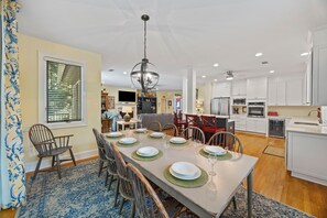 Une cuisine moderne avec des meubles blancs, un coin repas avec une table dressée pour quatre personnes et un salon doté d’une cheminée.