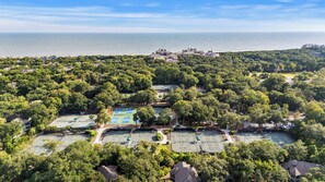 Une vue aérienne d’un quartier résidentiel avec de nombreux courts de tennis entourés d’une végétation luxuriante, une vue dégagée sur la mer et un ensemble de bâtiments situés au loin.