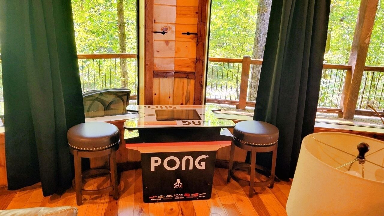Una mesa de Pong arcade con taburetes, una puerta de madera y vistas a árboles a través de amplias ventanas.