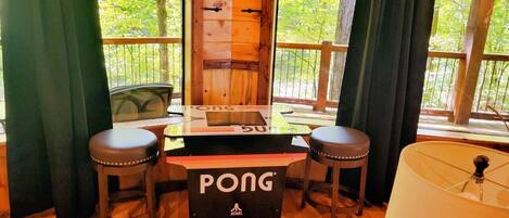 Una mesa de Pong arcade con taburetes, una puerta de madera y vistas a árboles a través de amplias ventanas.
