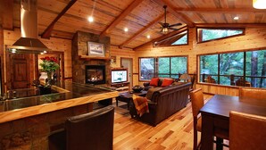 A wooden cabin interior with a stone fireplace, a dining area, and large windows offering a forest view.