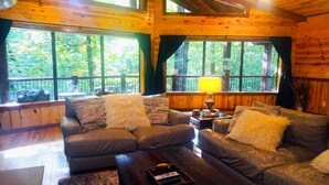 A cozy cabin interior with a wooden interior, two sofas with fluffy pillows, a coffee table, and large windows offering a view of the forest.