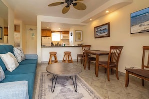 A living room with a blue sofa, two wooden chairs, a round table, and a kitchen in the background. - Kealia 407 Partial Ocean View Comfortable Charming Paradise Unit Washer/Dryer (Kihei)