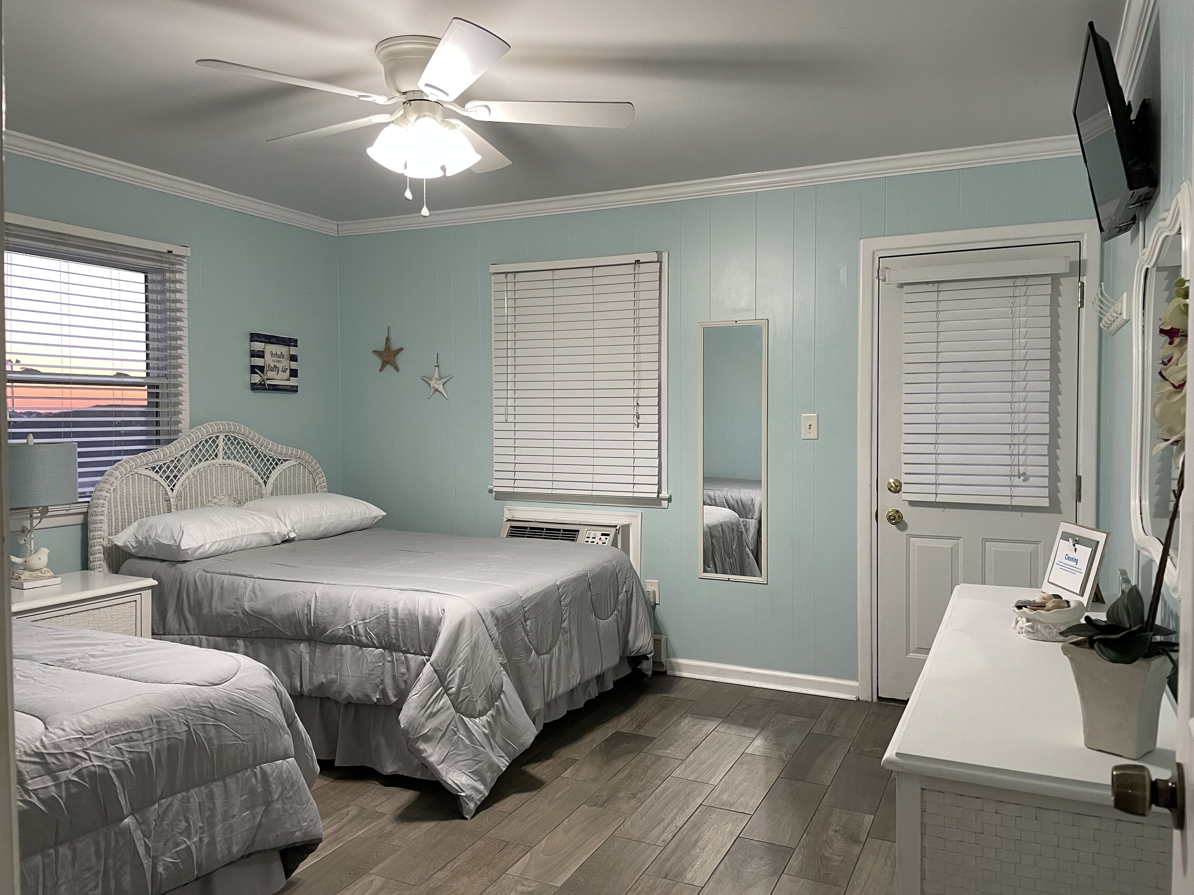 A bedroom with two beds, a ceiling fan, a window with blinds, a mirror, and a door with a sign.