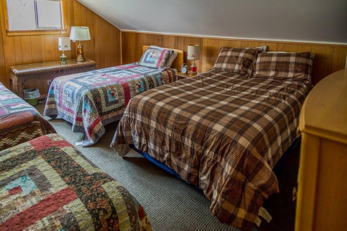Une pièce chaleureuse avec trois lits, chacun recouvert d’une couverture différente, des murs en bois et une petite table de chevet avec une lampe.
