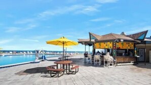 Ein Poolbereich mit Tischen, Sonnenschirmen, einer Bar und Blick auf den Ozean.