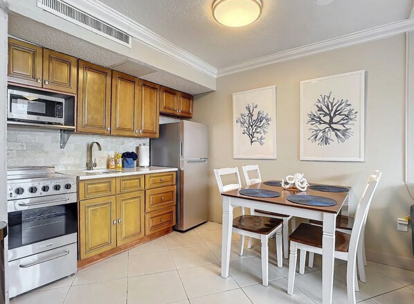 A kitchen with wooden cabinets, a built-in oven, a microwave, and a refrigerator. There is a dining table with chairs and two framed artworks on the wall.